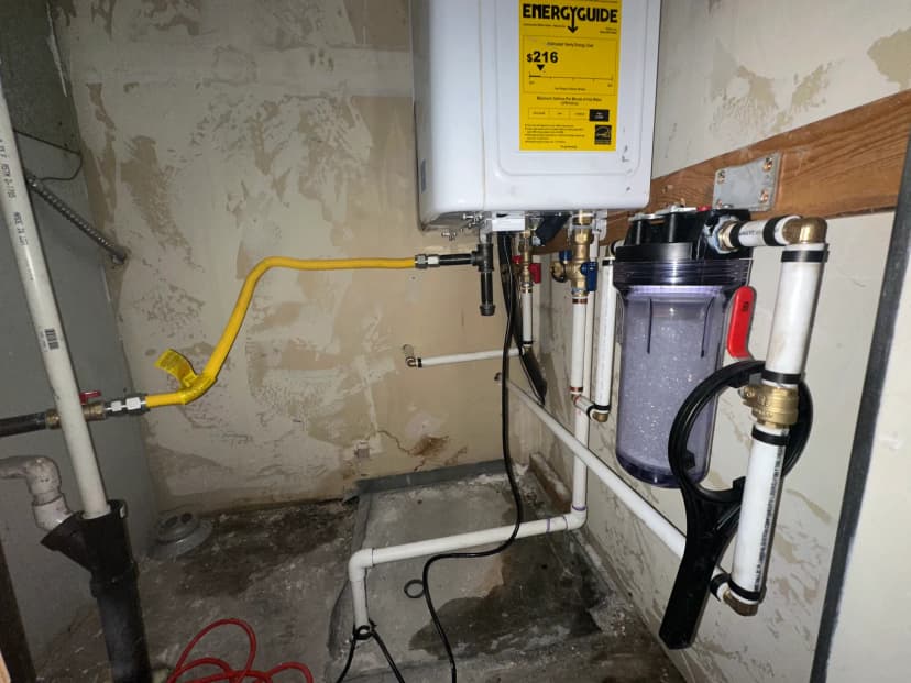 The underside of a wall-mounted tankless water heater with a yellow gas line, white water pipes, and a clear filter housing.