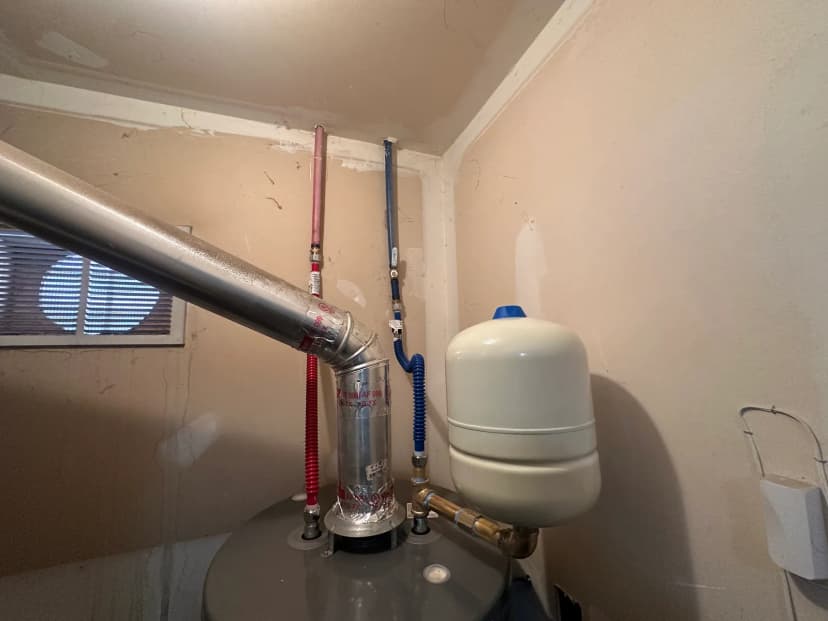 La parte superior de un calentador de agua con un tanque de expansión, líneas flexibles de agua y un tubo de ventilación metálico.