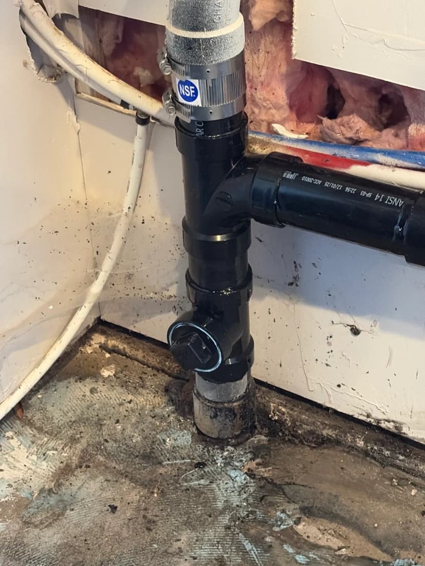 Un tubo de drenaje negro de ABS con registro de limpieza, unido a un tubo gris en una pared abierta.