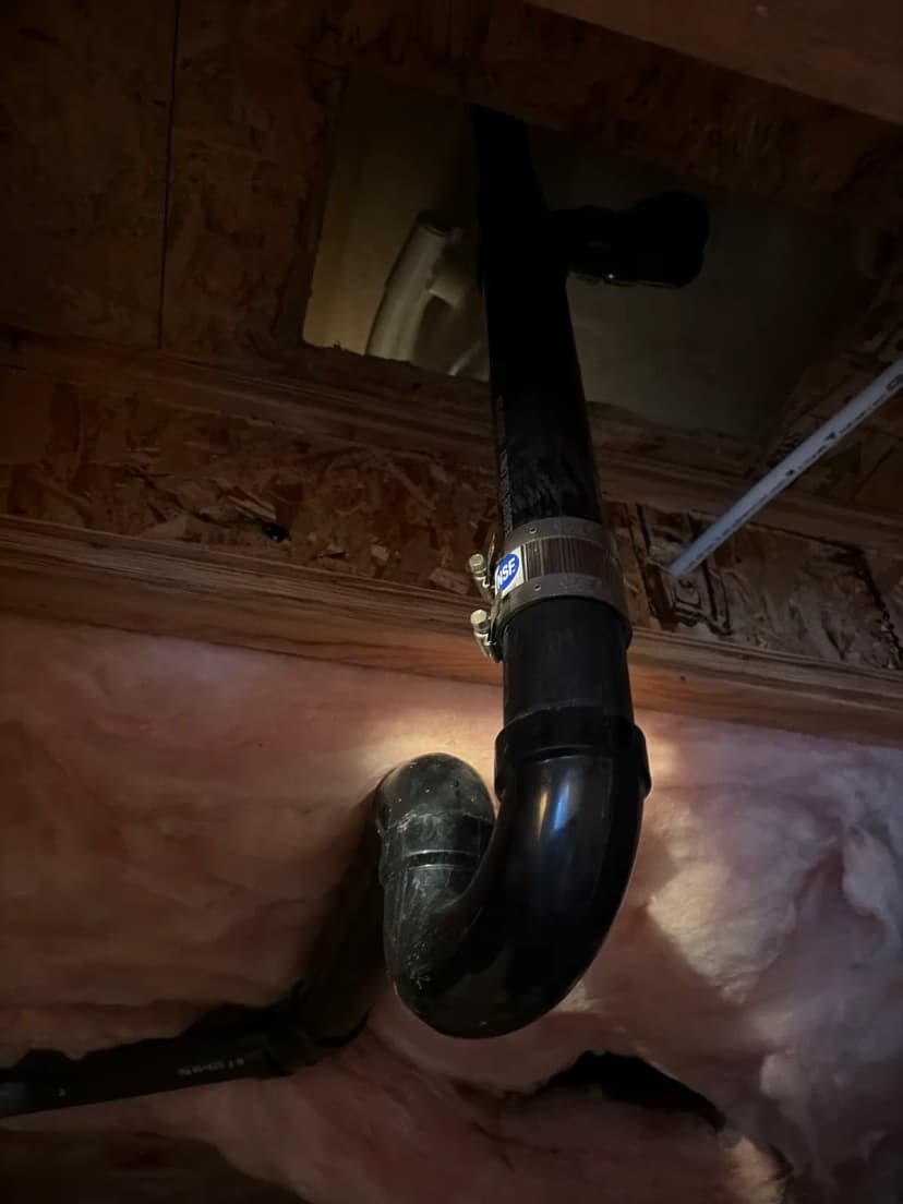 A black plastic drain pipe and trap seen from below, coming down through an opening in a wood subfloor past pink insulation.