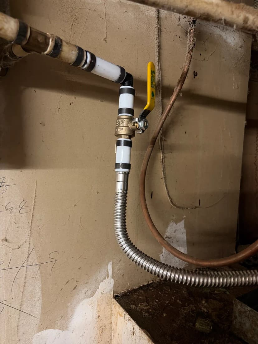 A brass ball valve with a yellow handle on white pipe, connected to a corrugated stainless steel water line.