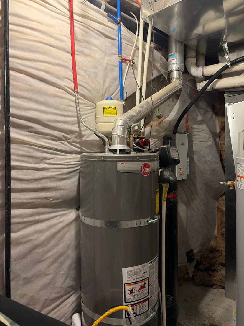A gray water heater strapped to a wall, with an expansion tank, flexible water lines, and a metal vent pipe above it.