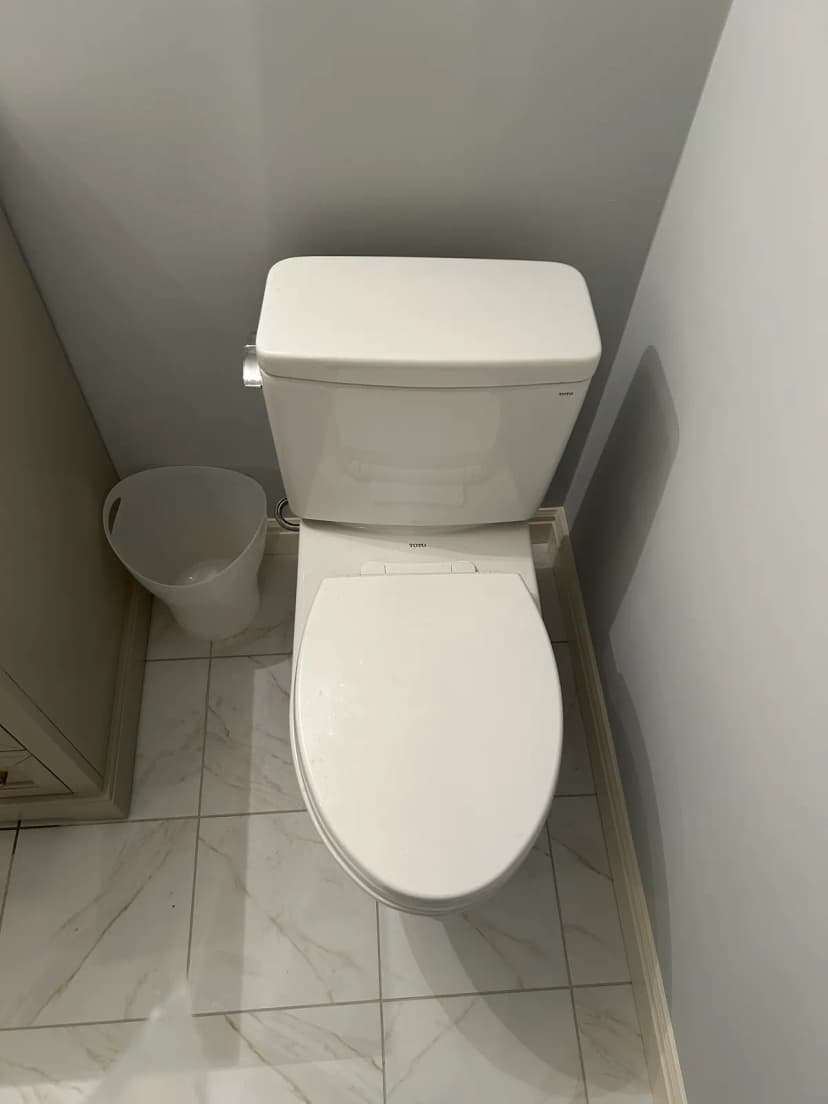A white two-piece toilet on a white tile floor in the corner of a bathroom, with a small wastebasket beside it.