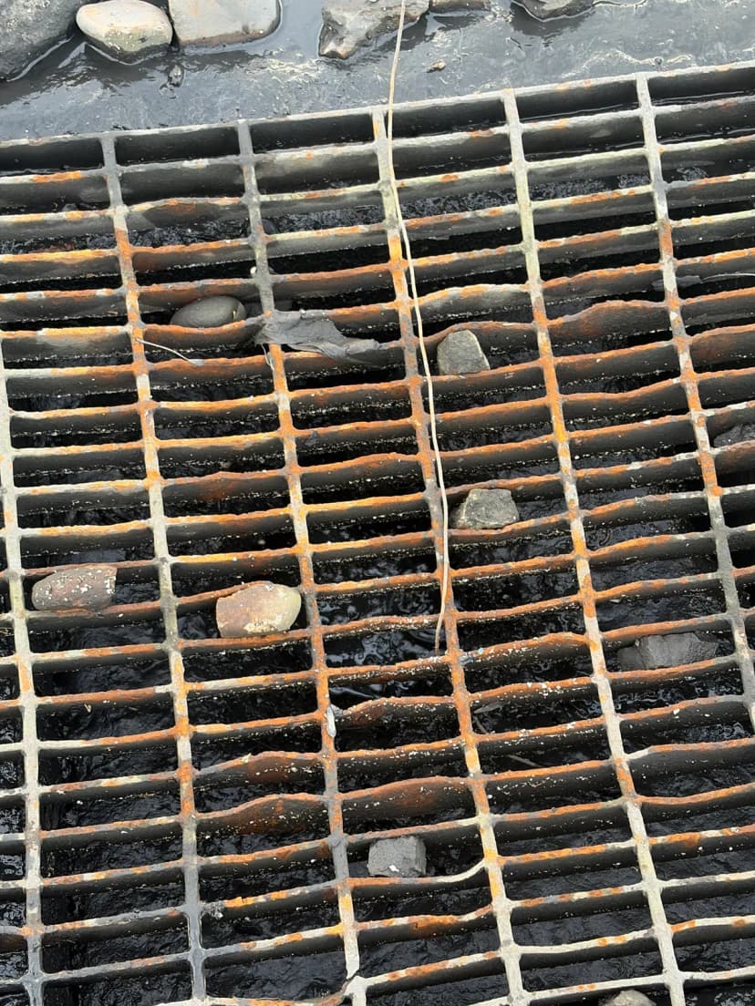 A rusted steel storm drain grate with rocks on it and water below.