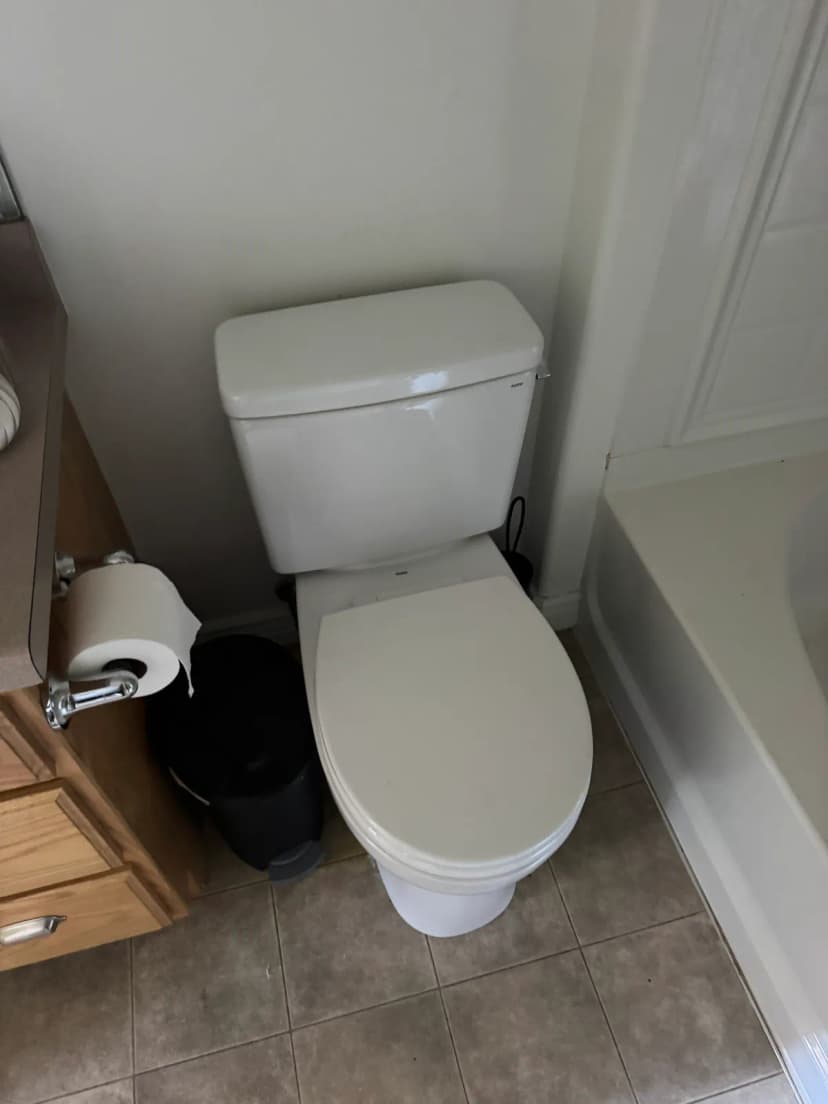 A white toilet with the lid closed, between a wood vanity and a bathtub, on a tile floor.