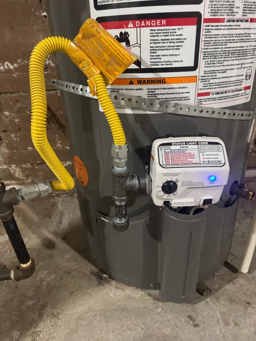 A yellow flexible gas line running into a tee with a capped pipe below it, at the gas control of a gray tank water heater.