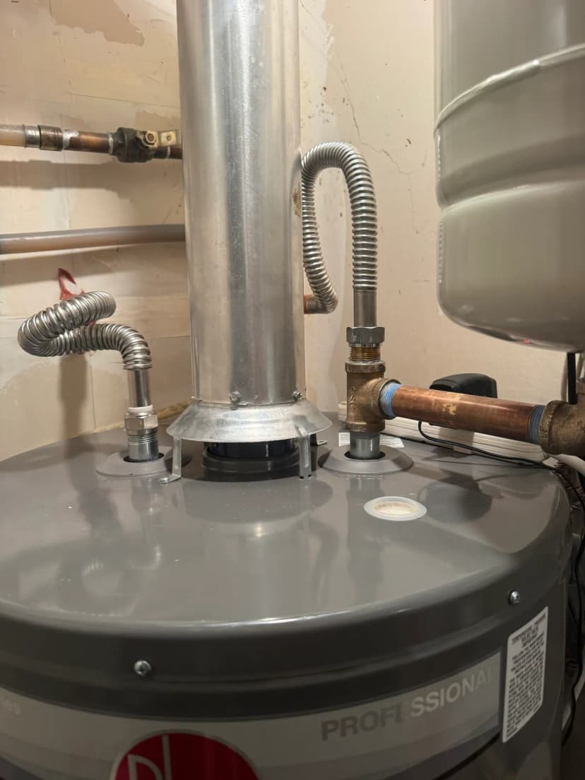 The top of a gray tank water heater with a metal vent pipe, two stainless flex lines, and an expansion tank beside it.