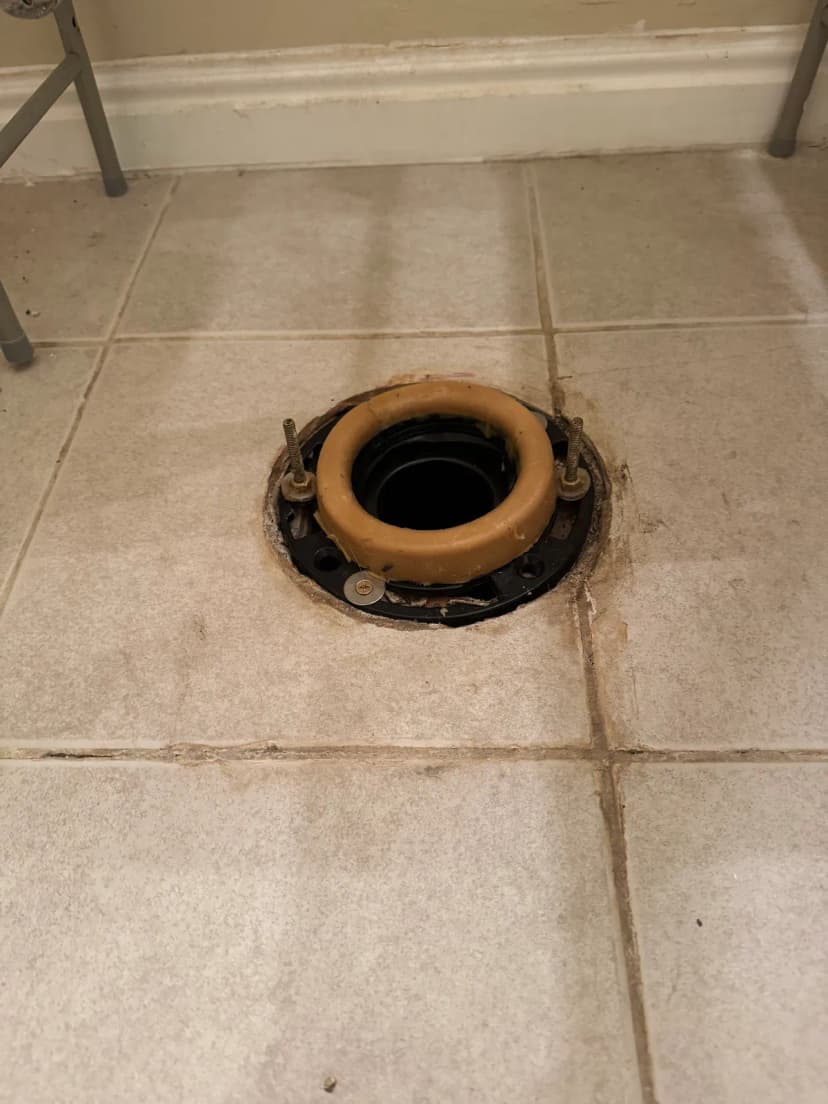 A black toilet flange with a wax ring and two bolts, set in a tile floor.