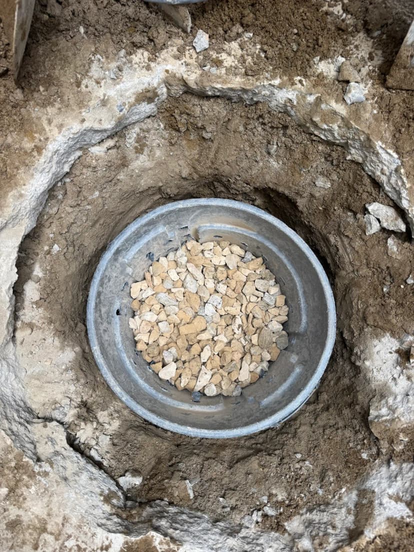 A metal drum filled with gravel, set in a hole dug through concrete.