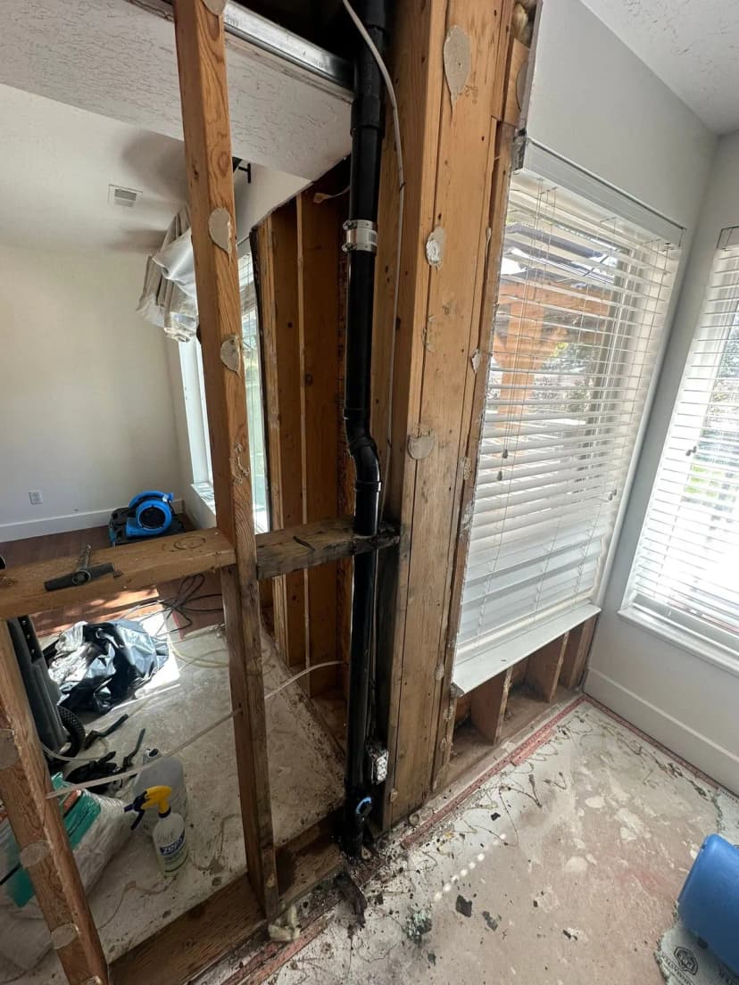 A black drain pipe running down an opened wall between wood studs, beside a window.