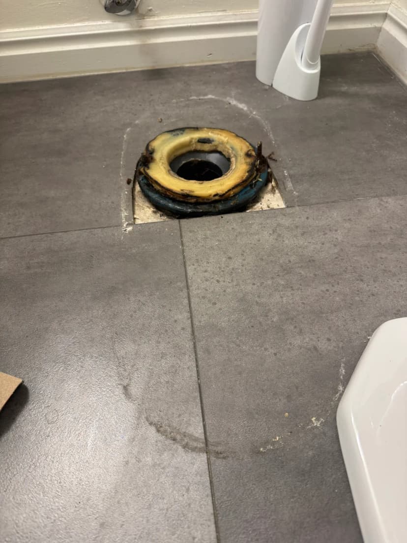 A toilet flange with an old wax ring in a tile floor, with the toilet removed.