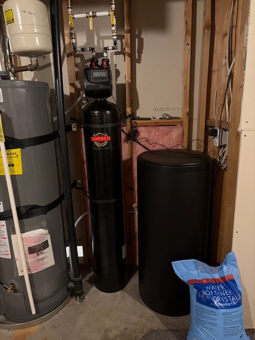 A water softener tank and a brine tank beside a water heater, with a bag of softener salt on the floor.